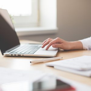 computer sitting on desk with woman's hand touching the computer and a notebook and pen next to the computer on a desk