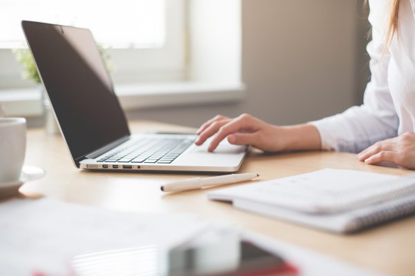 computer sitting on desk with woman's hand touching the computer and a notebook and pen next to the computer on a desk